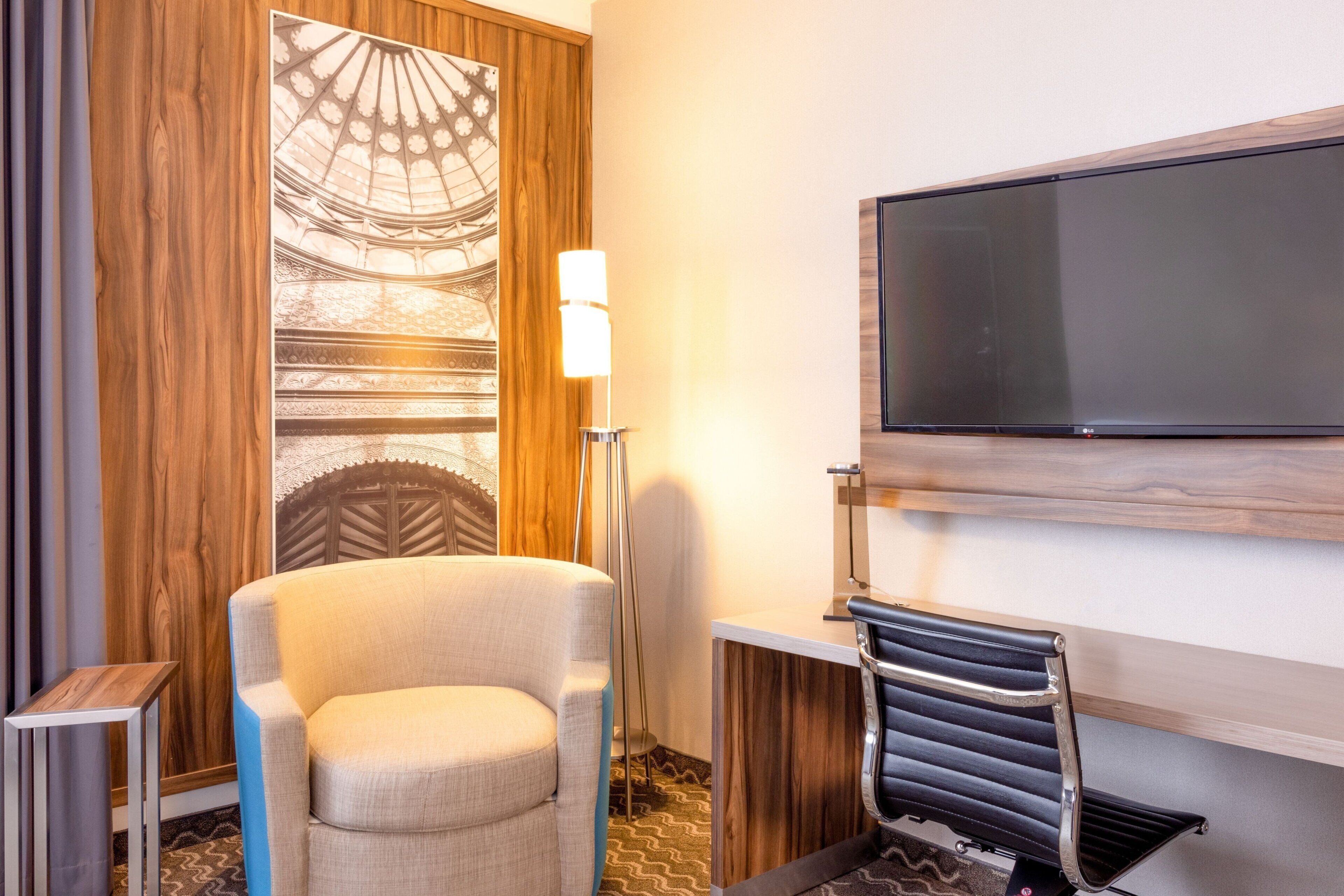 A hotel room with a large flat-screen TV mounted on the wall, a comfortable armchair, a standing lamp, and a radiator.