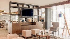 Un moderno vestíbulo de hotel con un televisor de pantalla plana grande, asientos cómodos, un mostrador de recepción de madera y un ventanal con vistas a la ciudad.