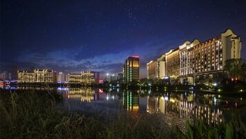 Vista nocturna de un hotel con varios edificios altos, un lago tranquilo que refleja las luces y un cielo estrellado.
