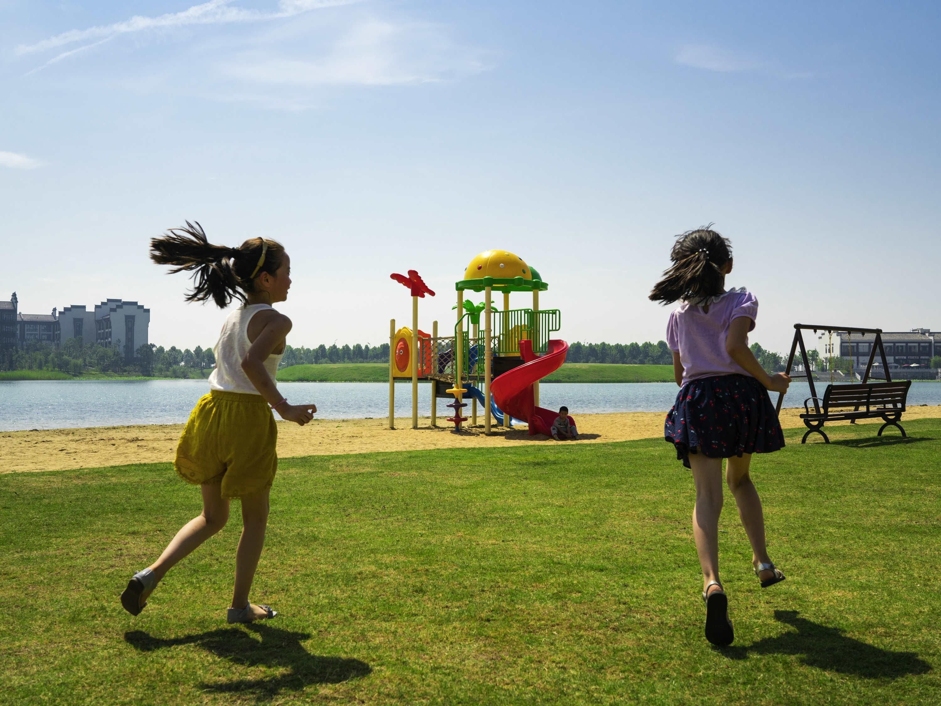 Dos niños jugando cerca de un parque infantil con un lago y edificios al fondo.