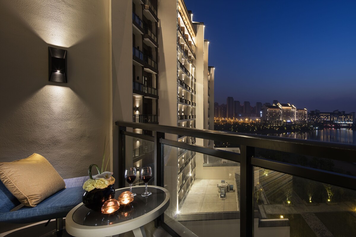 A balcony with a sofa, a table with a vase and glasses, and a city view at dusk.