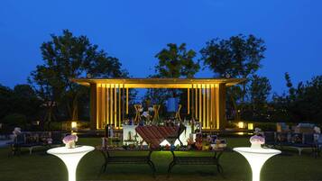 An outdoor event space with a stage, tables, and chairs, surrounded by trees.