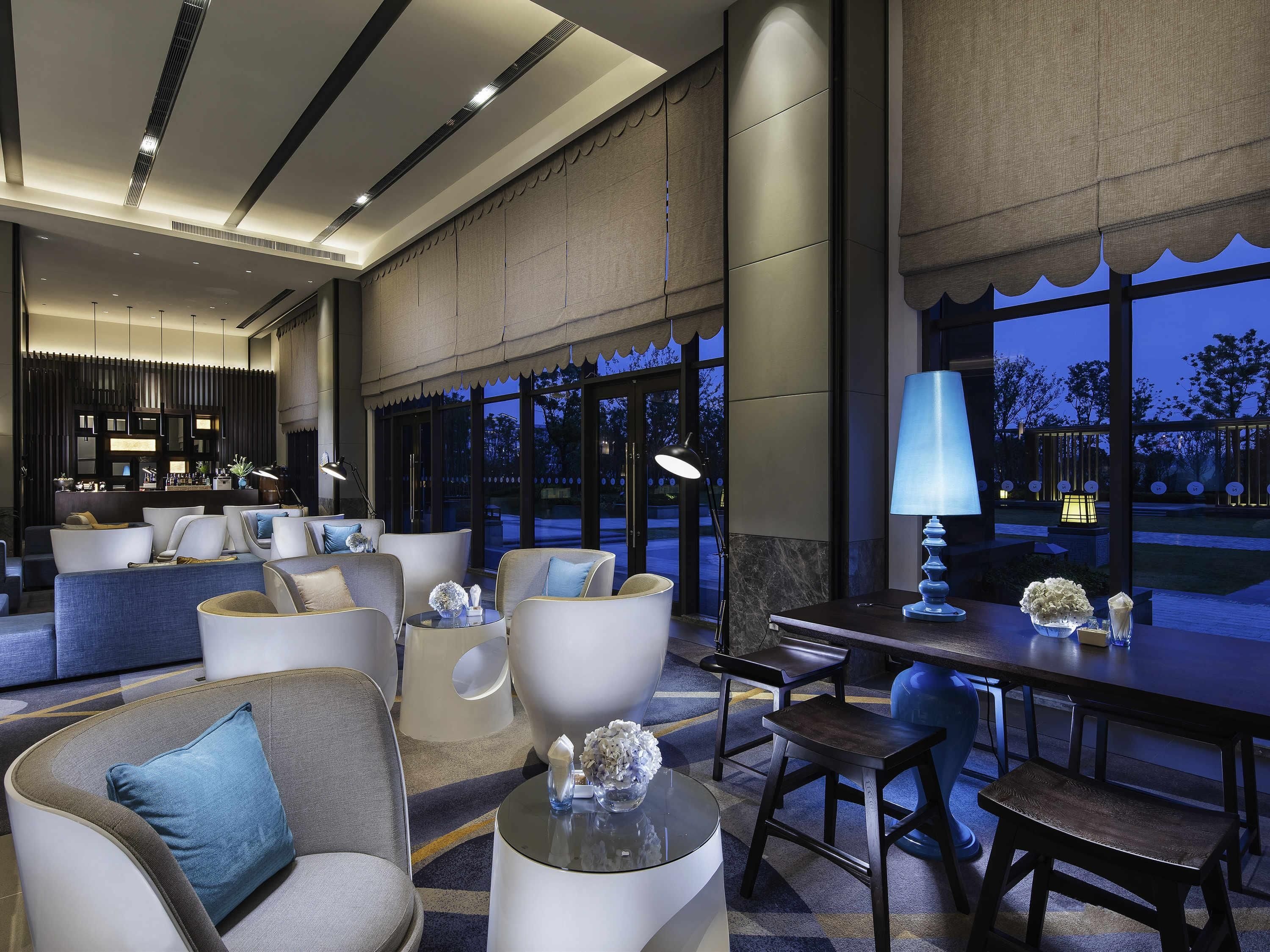 A modern hotel lobby with seating areas, a bar, and large windows offering views of the outdoors.