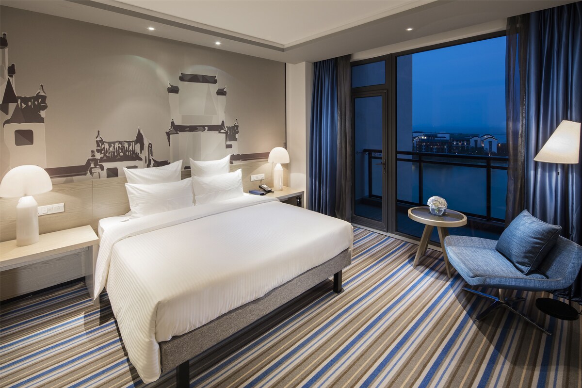 A hotel room with a large bed, a seating area, a small table, and a view of the cityscape through the window.
