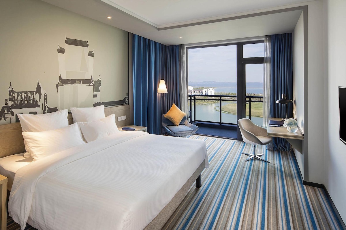 Habitación de hotel con una cama grande, un escritorio con silla, un televisor y vista a un cuerpo de agua.