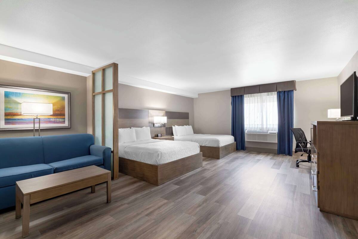 A hotel room with a blue sofa, two single beds, a wooden desk, a TV, and a painting on the wall.