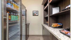 Een smalle gang met een koelkast vol drankjes, een magnetron en een kleine koelkast. Er staan planken met snacks en een lijst met gerechten aan de muur.