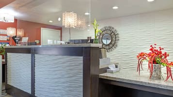 Uma área de recepção com design moderno, com um balcão de atendimento, um espelho decorativo e um vaso com flores vermelhas.