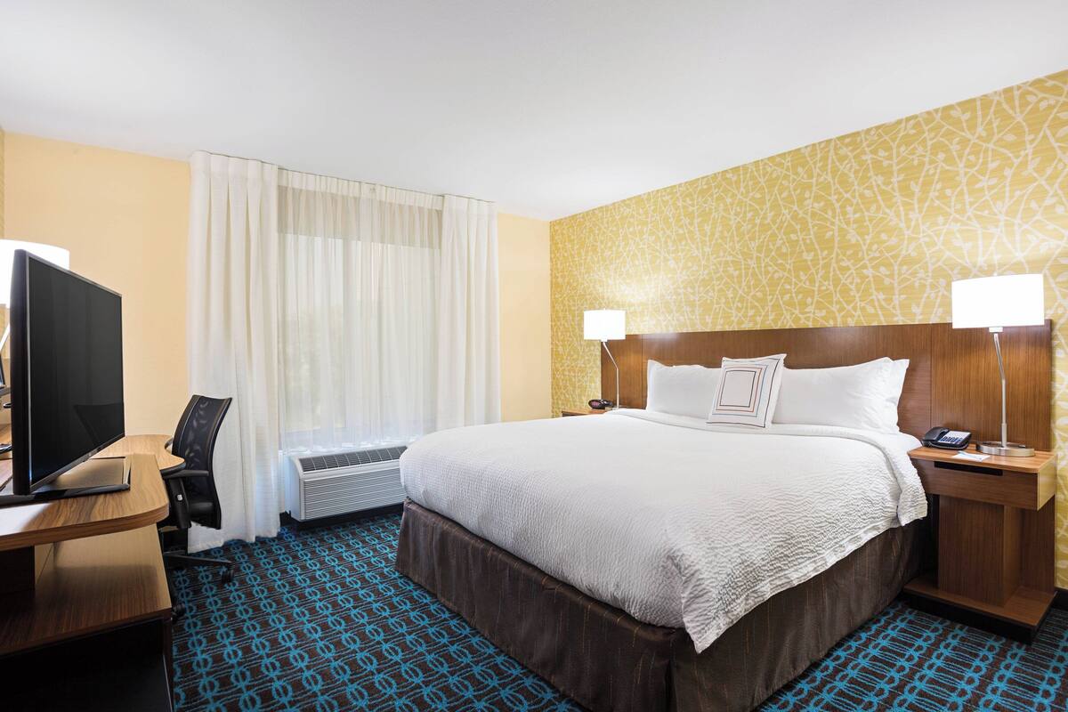 A hotel room with a bed, a desk with a computer, a TV, and a window with curtains.