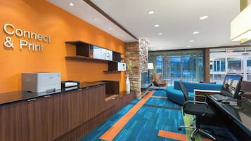 Zona de recepción con un letrero que dice 'Connect & Print', una impresora Samsung, un pilar de piedra, un sofá azul y un escritorio con una computadora portátil.
