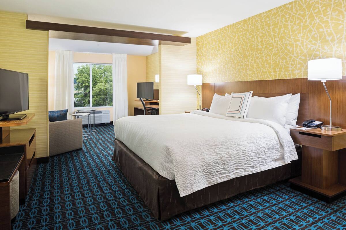 A hotel room with a large bed, a desk with a computer, a TV, and a window with curtains.
