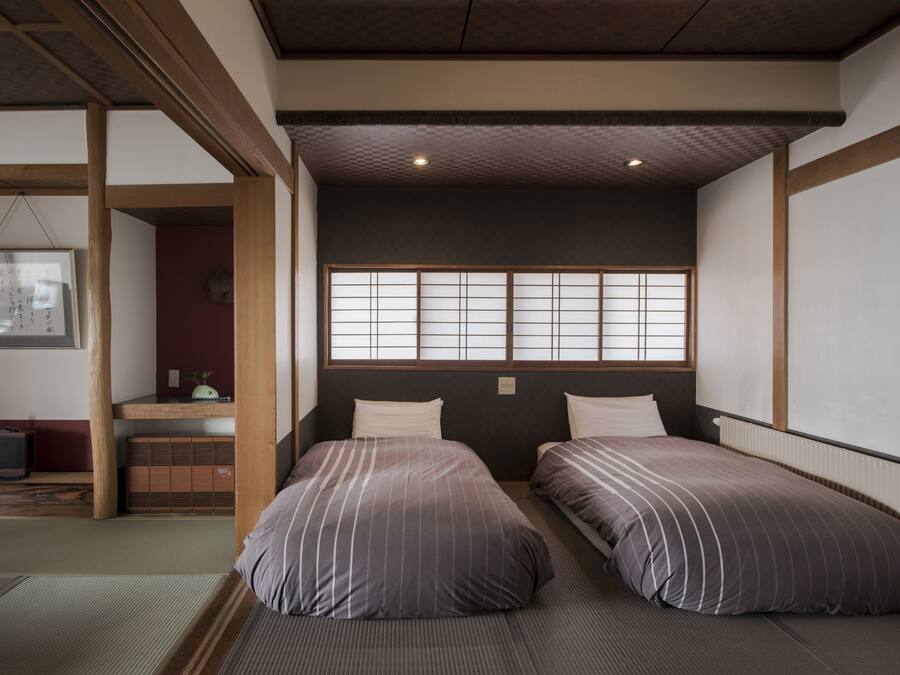 A traditional Japanese room with tatami flooring, two beds with striped bedding, a shoji screen window, and a small desk with a chair.