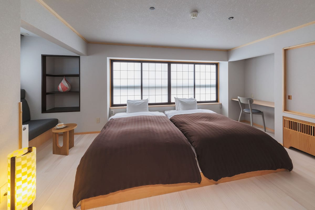 Ein modernes Schlafzimmer mit großem Fenster, einem Bett mit dunkler Bettwäsche, einem Schreibtisch und einem Stuhl.