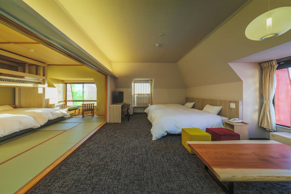 A hotel room with two beds, a wooden table, a small desk, and a window with curtains.