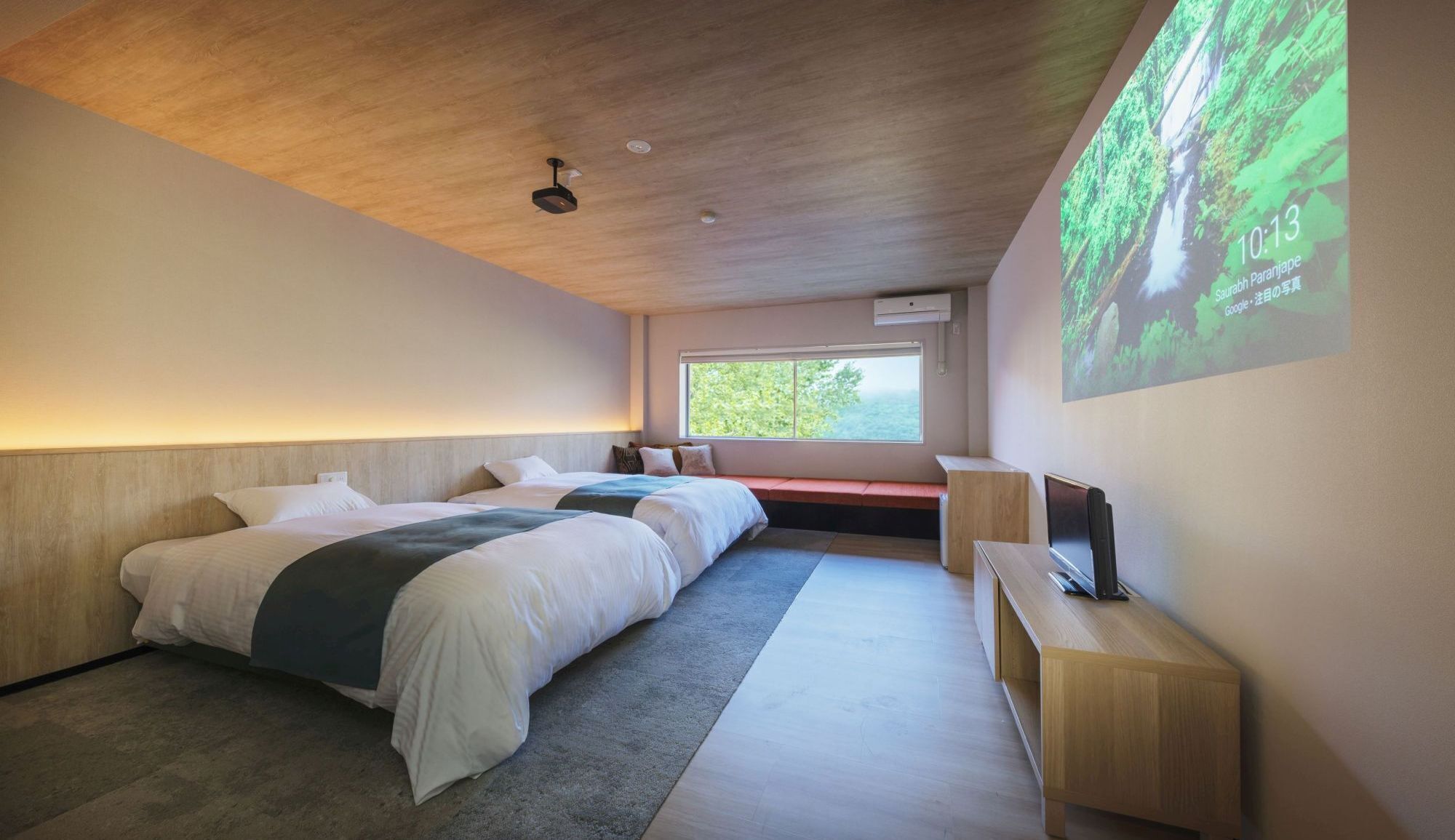 Une chambre d’hôtel moderne avec deux lits, une grande fenêtre, un écran de projection et un plafond en bois.