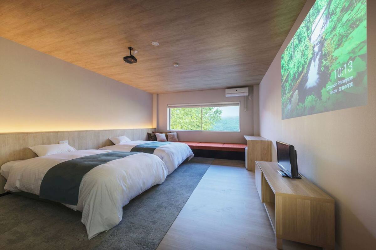 A modern hotel room with two beds, a large window, a projector screen, and a wooden ceiling.