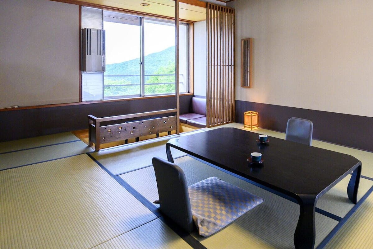 A traditional tatami room with a low table, chairs, a bench, and a view of a mountain.