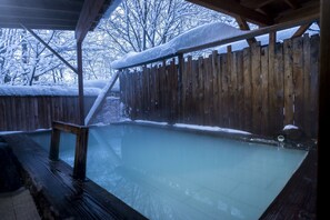 Une terrasse en bois avec un jacuzzi, entourée d’une clôture en bois et d’arbres couverts de neige.