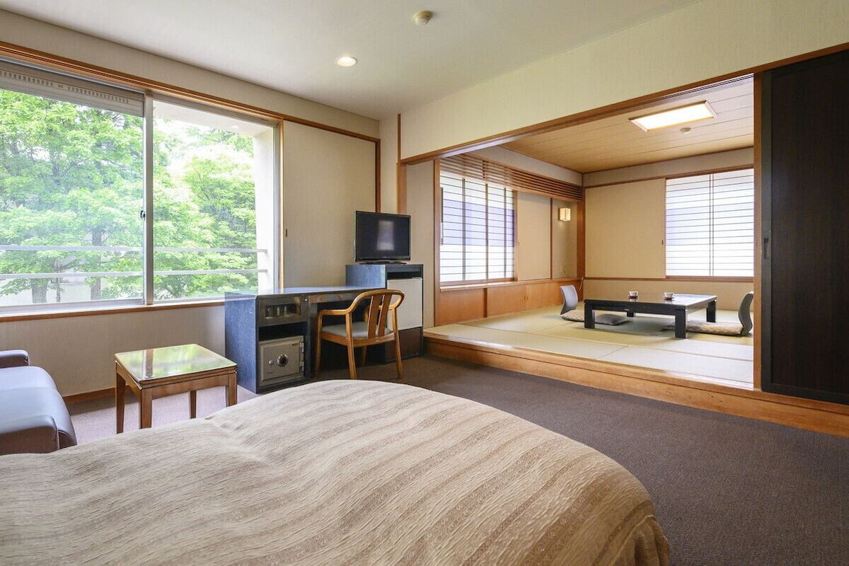 A hotel room with a bed, a desk with a television, a chair, a small table, and a view of trees through large windows.