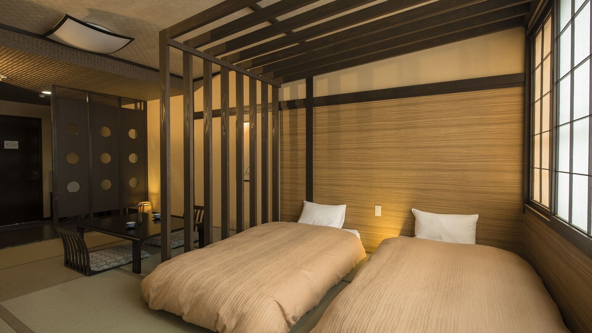 Ein traditioneller japanischer Raum mit Tatami-Boden, einem niedrigen Holzbett mit Kissen, einer Schiebetür mit runden Ausschnitten und Blick ins Freie durch ein Fenster.