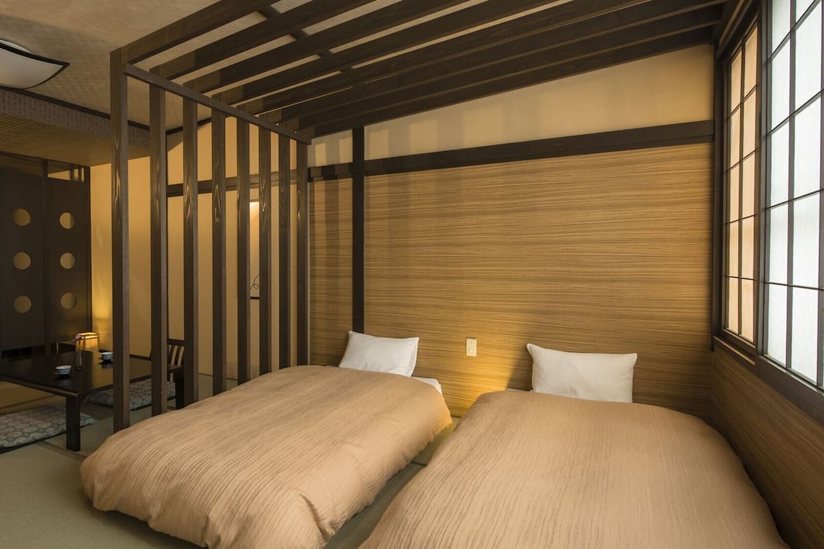 A traditional Japanese-style room with tatami flooring, a low wooden bed with pillows, a sliding door with circular cutouts, and a view of the outside through a window.