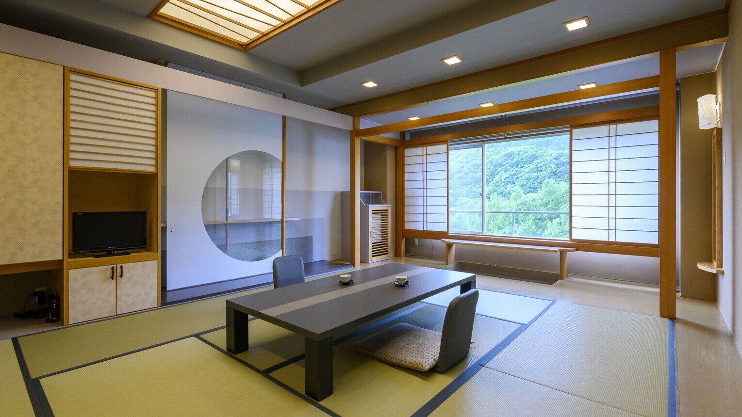 Ein traditioneller japanischer Raum mit Tatami-Boden, einem niedrigen Tisch, einem Fernseher und großen Fenstern mit Blick auf Grünflächen.