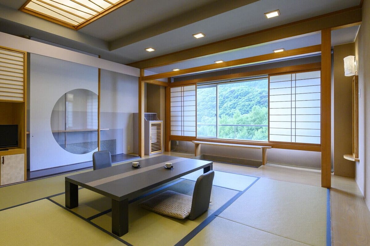 A traditional Japanese room with tatami flooring, a low table, a television, and large windows with a view of greenery.