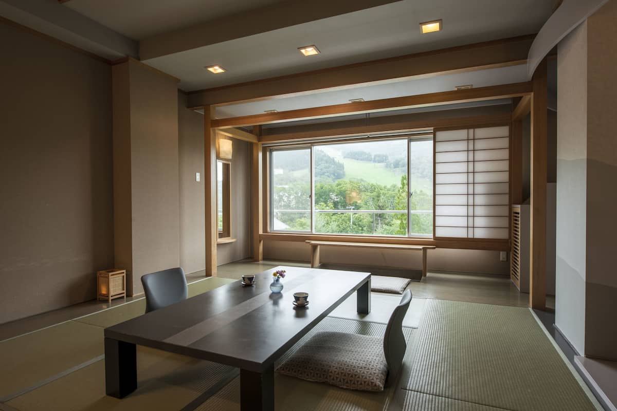 A traditional Japanese room with tatami flooring, a low table, and a view of a green landscape through a large window.