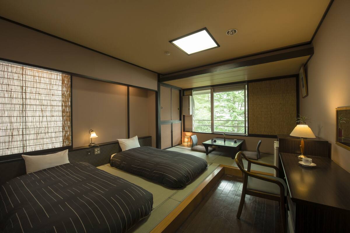 A hotel room with two beds, a desk, a chair, and a window with a view of trees.