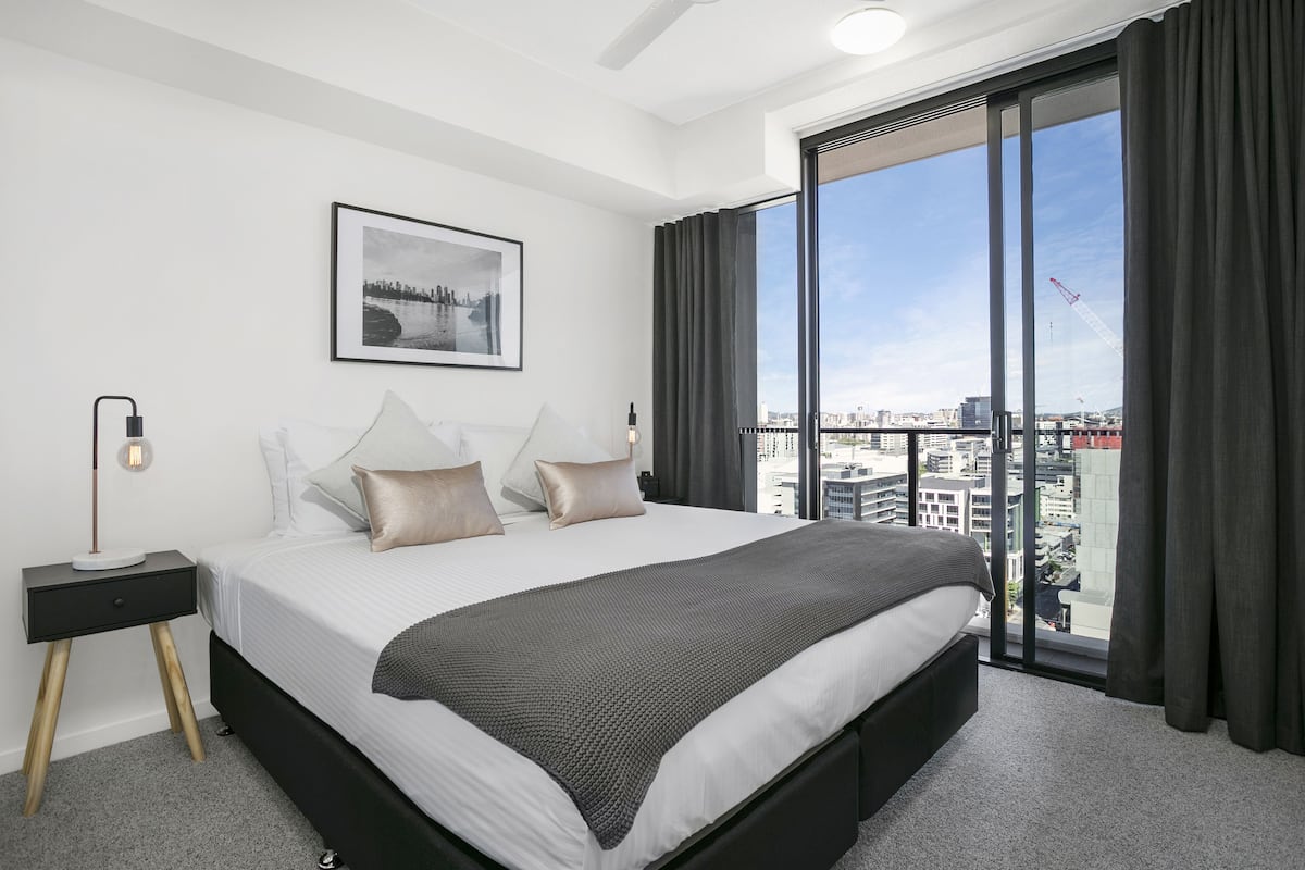 A modern hotel room with a large bed, a nightstand, a framed cityscape picture, and a view of the city through a large window.