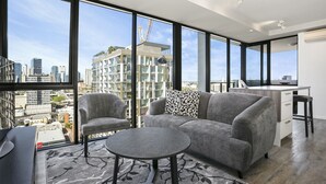 A modern living room with a gray sofa, two armchairs, a round coffee table, and a view of a cityscape through large windows. - Soda Apartments by CLLIX (South Brisbane)
