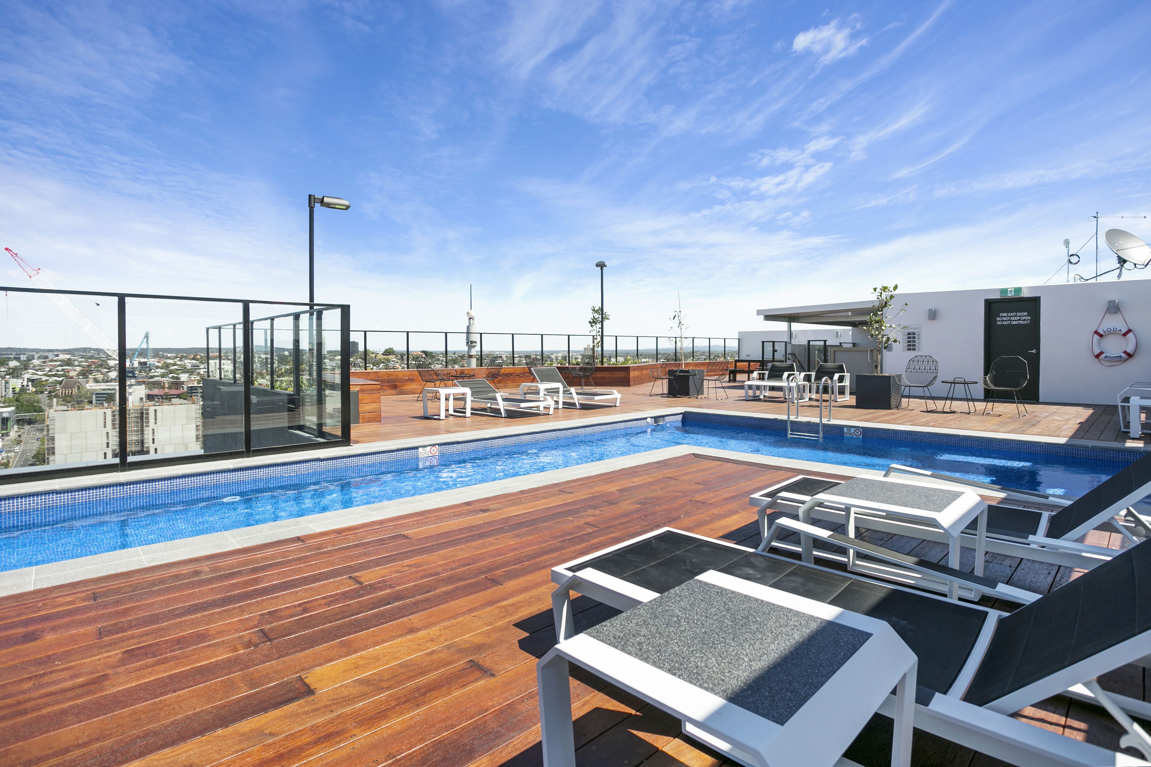 A rooftop pool with wooden decking and lounge chairs, surrounded by glass walls offering city views.