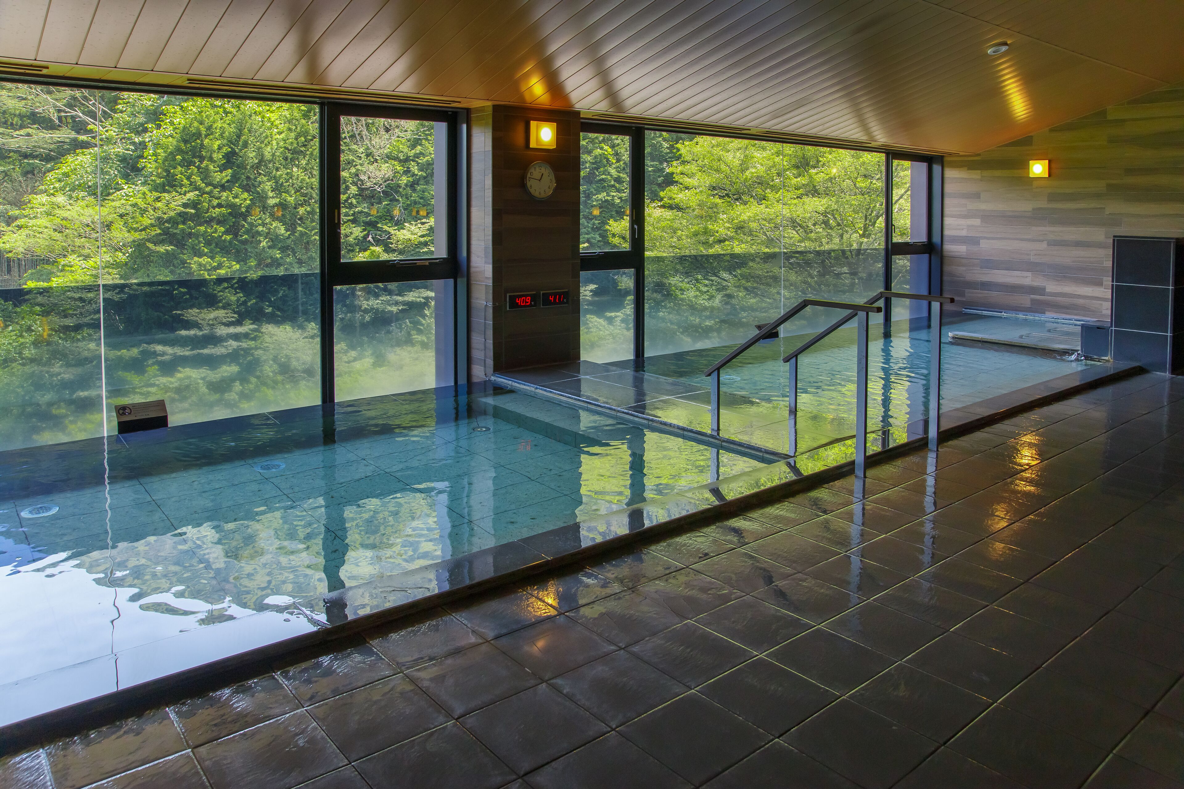 A modern indoor swimming pool with glass walls, a wooden ceiling, and a digital clock.