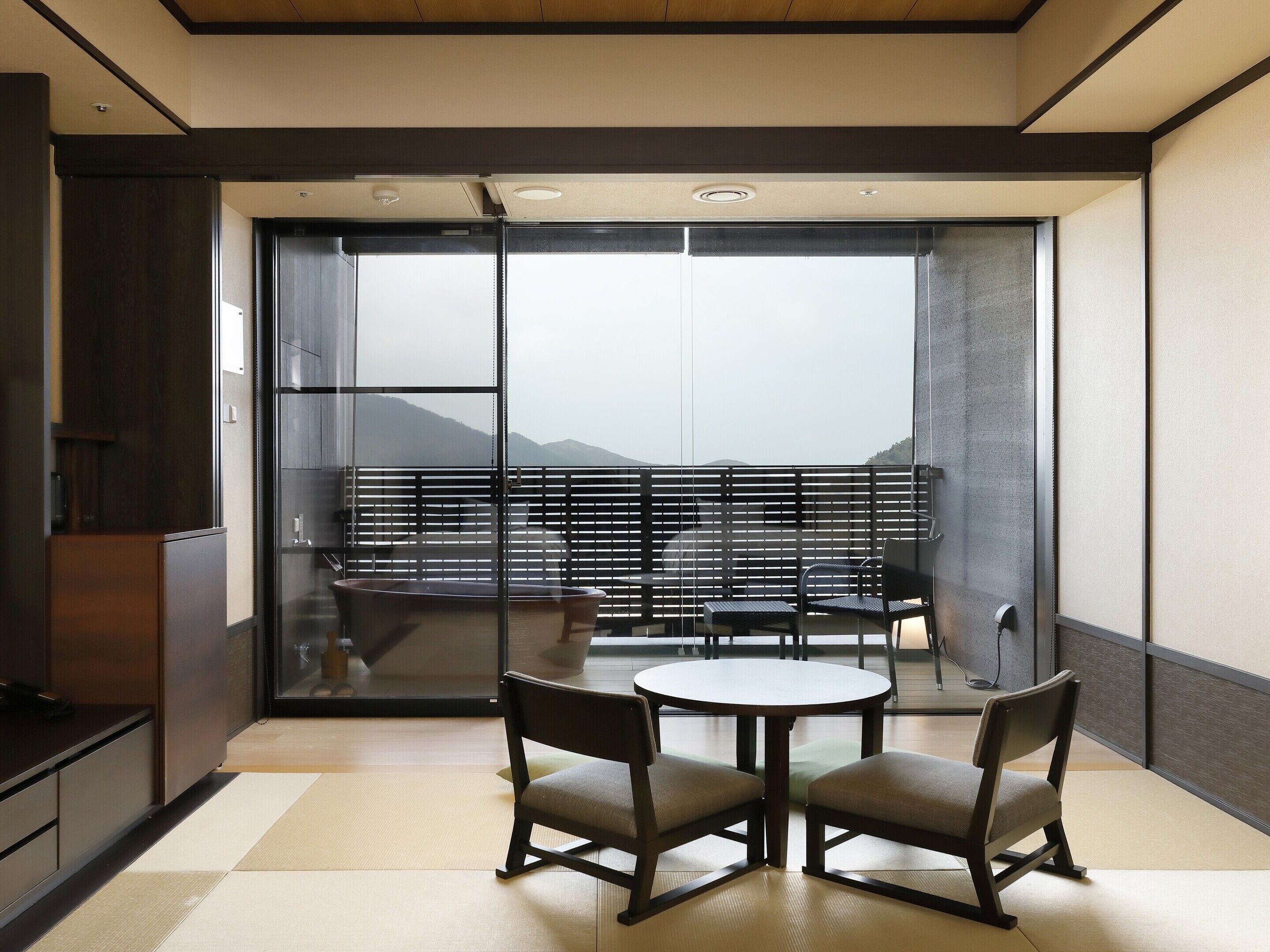 A modern room with a sliding glass door leading to a balcony with a view of mountains. There is a round table with two chairs and a bench with a view.