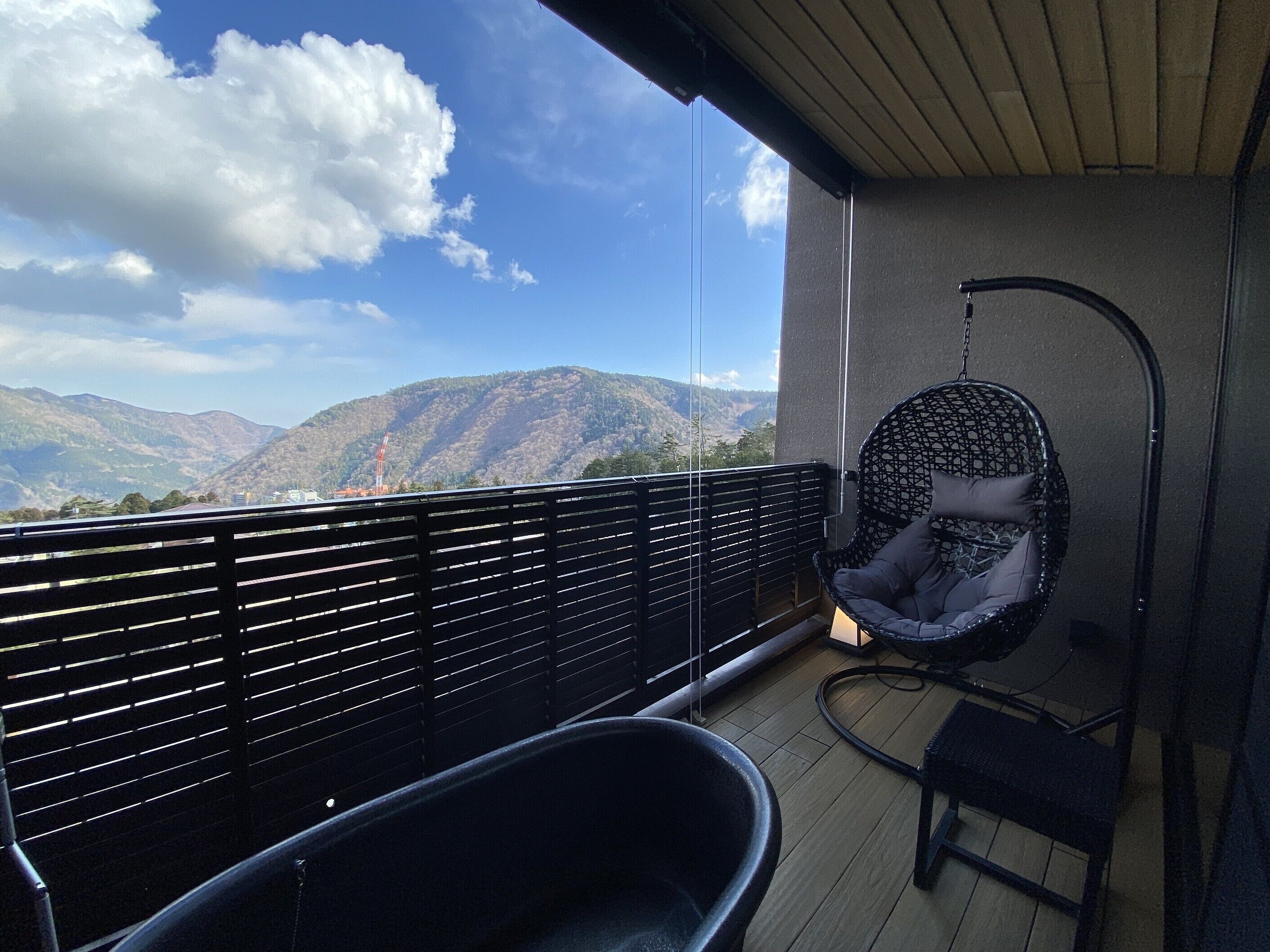 A balcony with a hanging chair and a black lounge chair, overlooking a mountainous landscape.