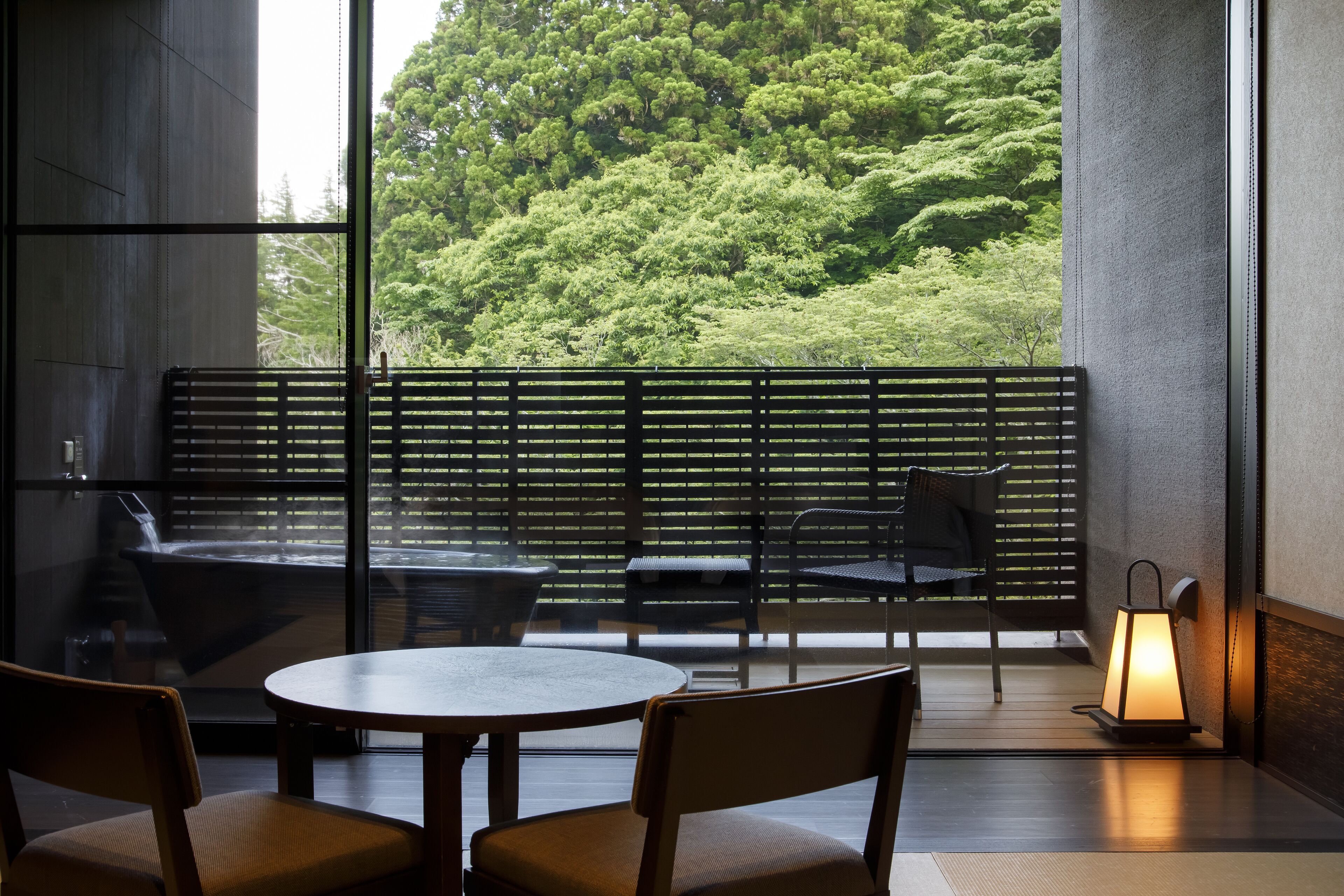 A modern interior with a round table and chairs, a bench, and a view of lush greenery through large windows.