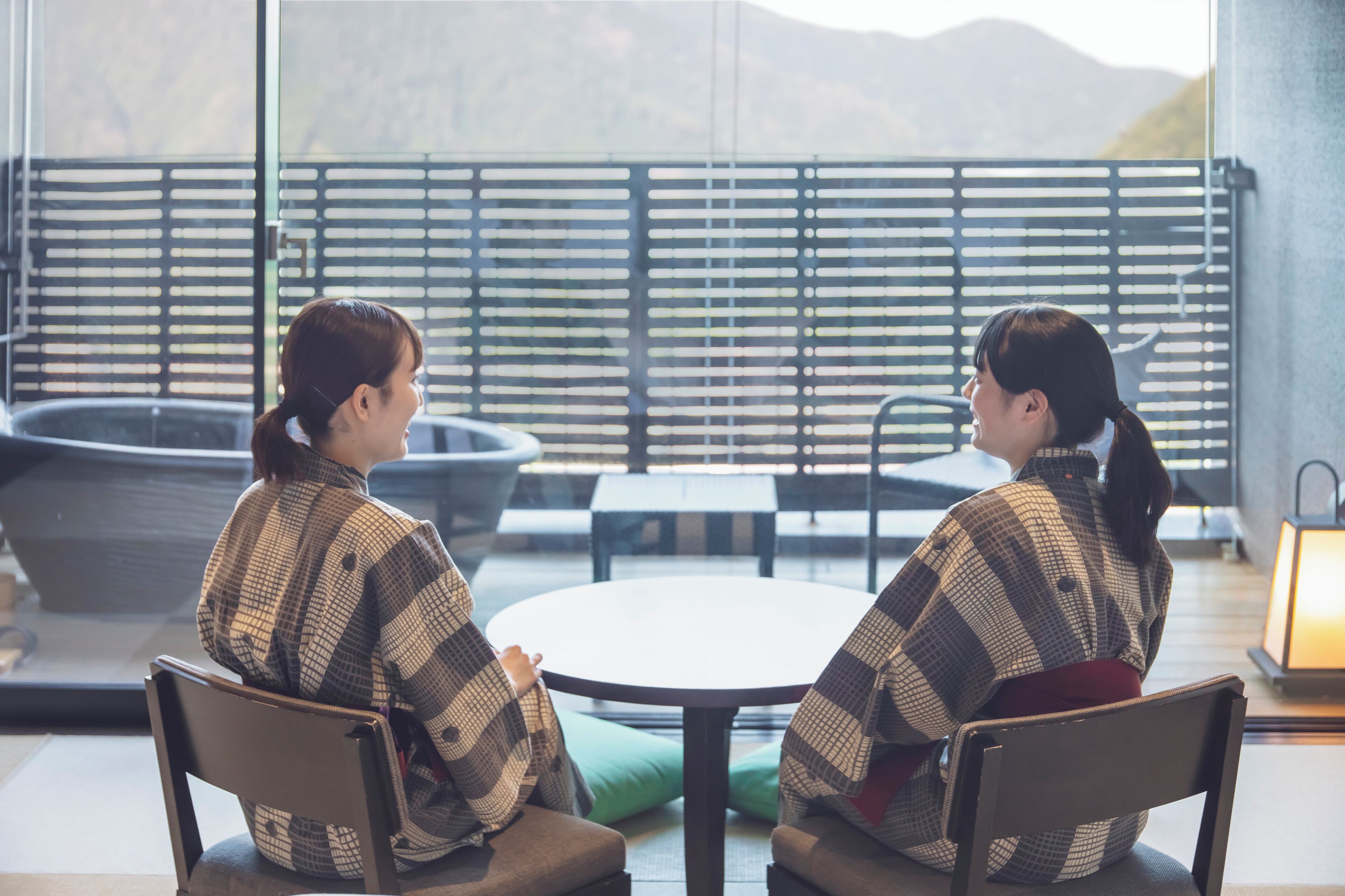 Two individuals seated at a round table in a modern interior with a view of mountains through a patterned window.