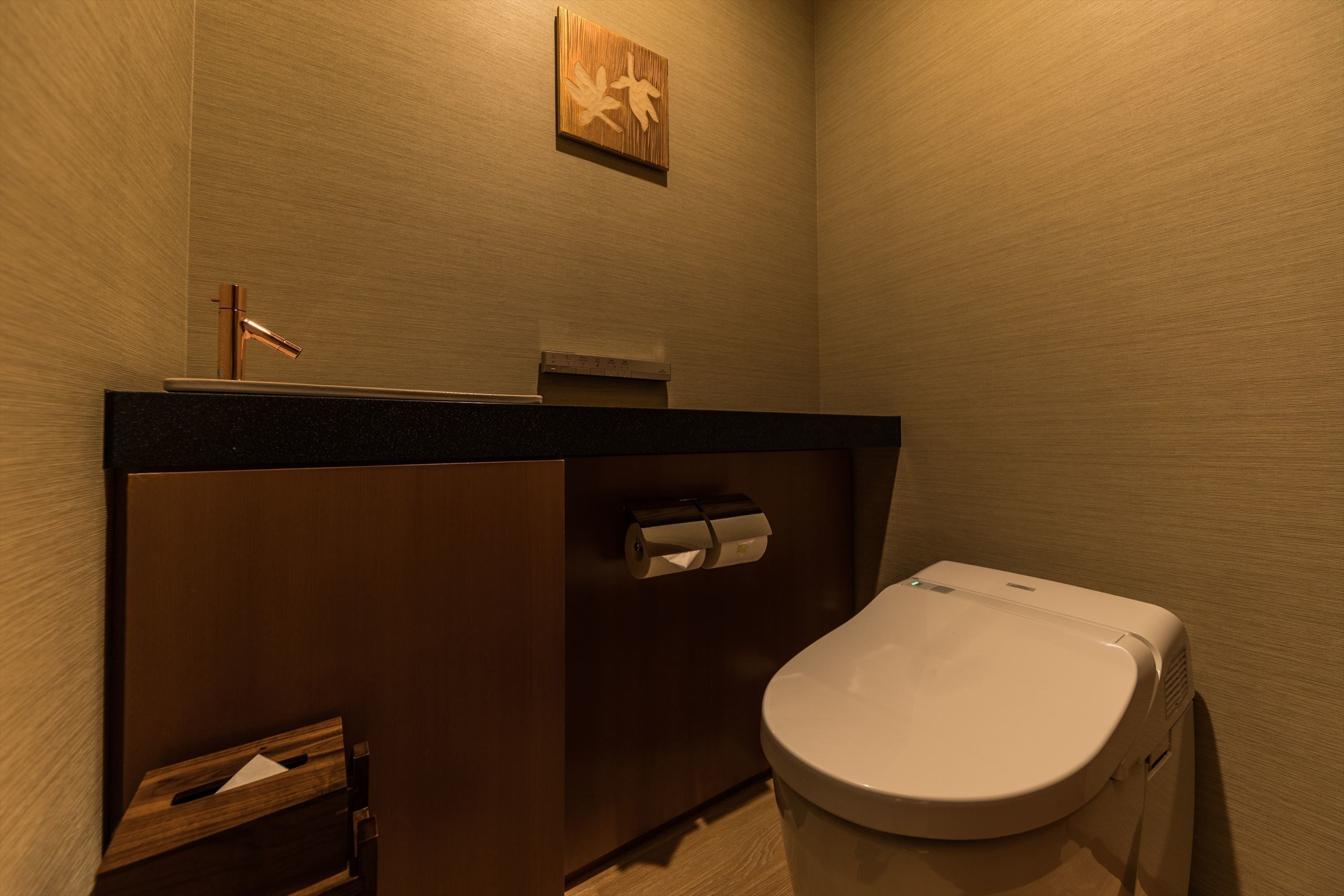 A compact bathroom with a wooden vanity, a toilet, and a paper towel dispenser.
