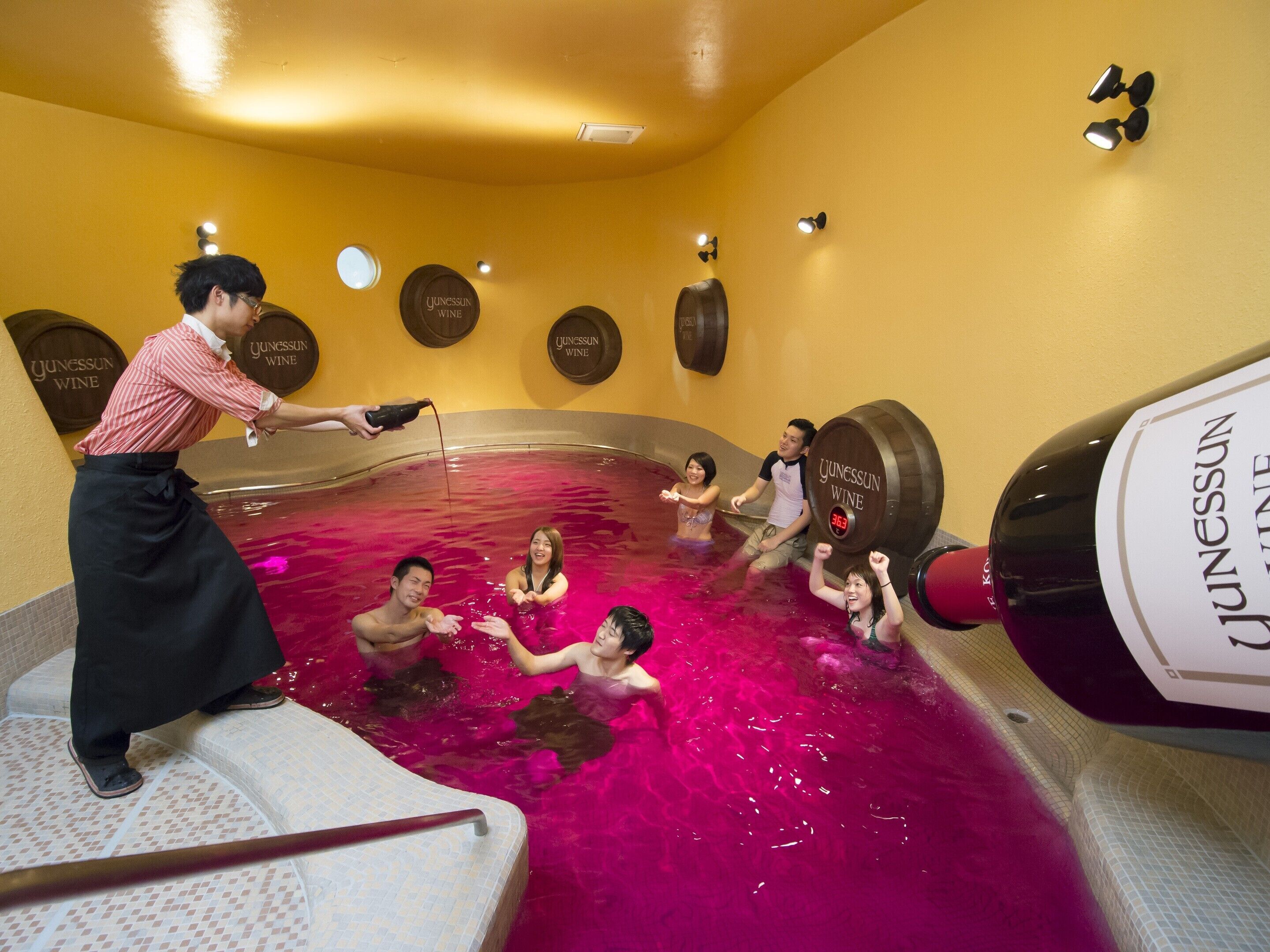 A group of people in a hot tub with a bartender pouring a drink. The room has a yellow wall and several round signs with 'Junessun Wine' written on them.