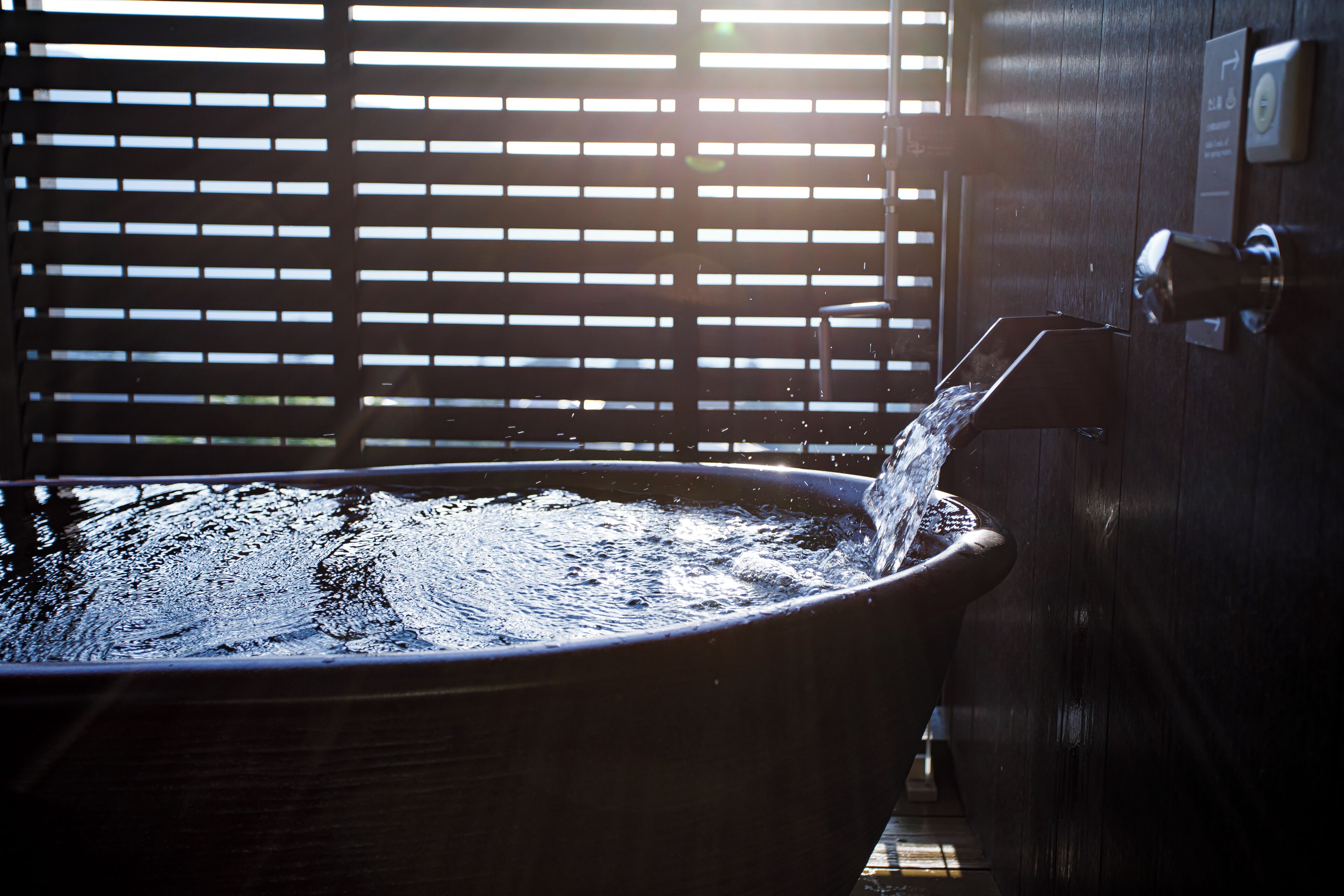 A large bathtub with water flowing into it, positioned next to a wooden door with a lock and a sign indicating a 24-hour service.