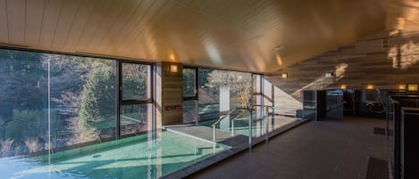 A modern indoor swimming pool with glass walls, wooden ceiling, and a view of the surrounding forest.
