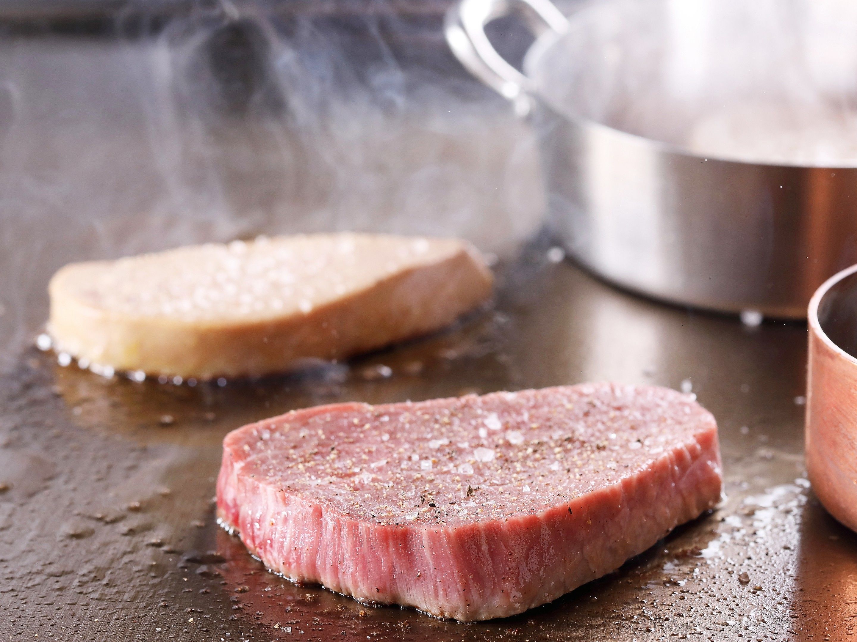 Two steaks on a grill with a pot in the background.