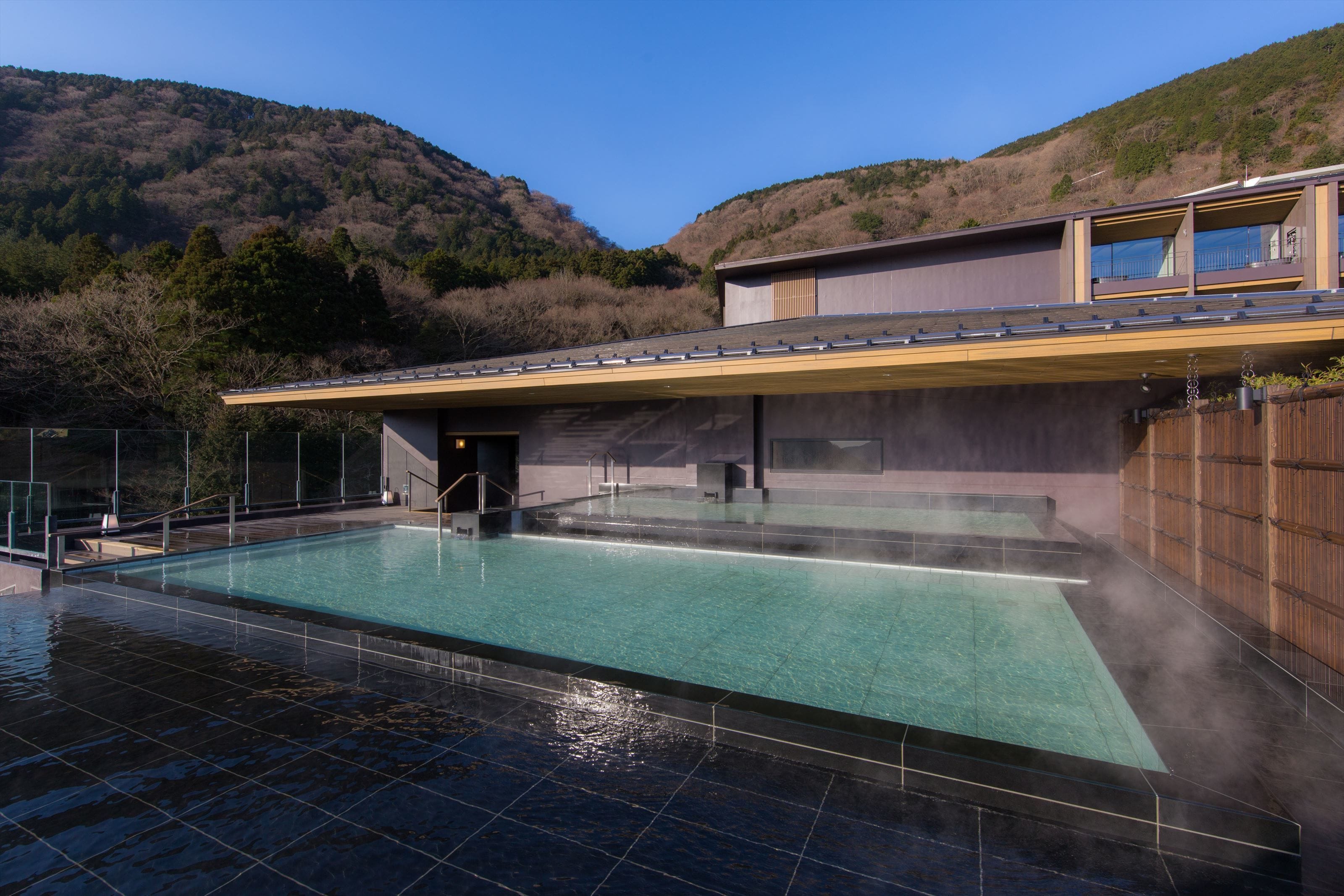 Un edificio moderno con piscina, rodeado por una valla de madera y con montañas al fondo.