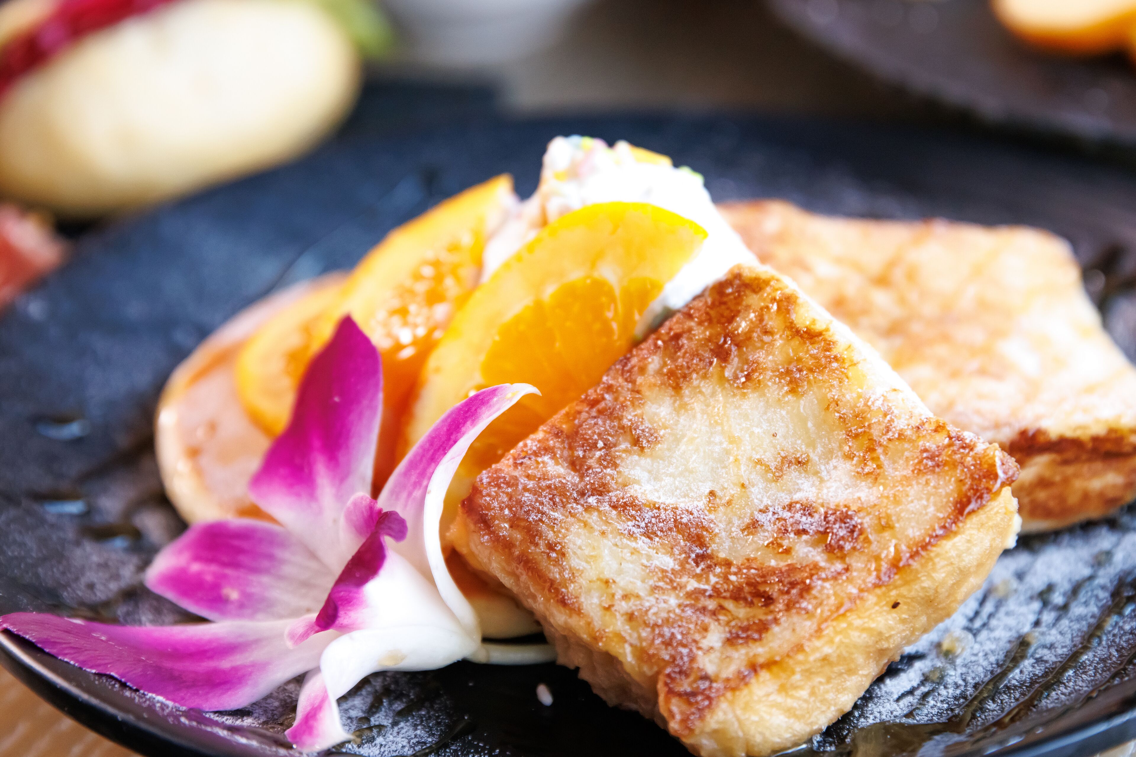 Primer plano de un postre con una rebanada de tostada, rodajas de naranja y una flor morada sobre un plato oscuro.