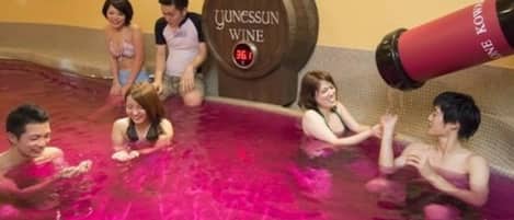 A group of people in a spa pool with a wooden barrel labeled "JUNESSUN WINE" and a red bottle with "NE KOWA" text.