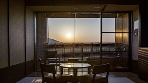 A room with a round table and two chairs, a bed with a patterned bedspread, and a view of the sun setting over a mountain through a large window.