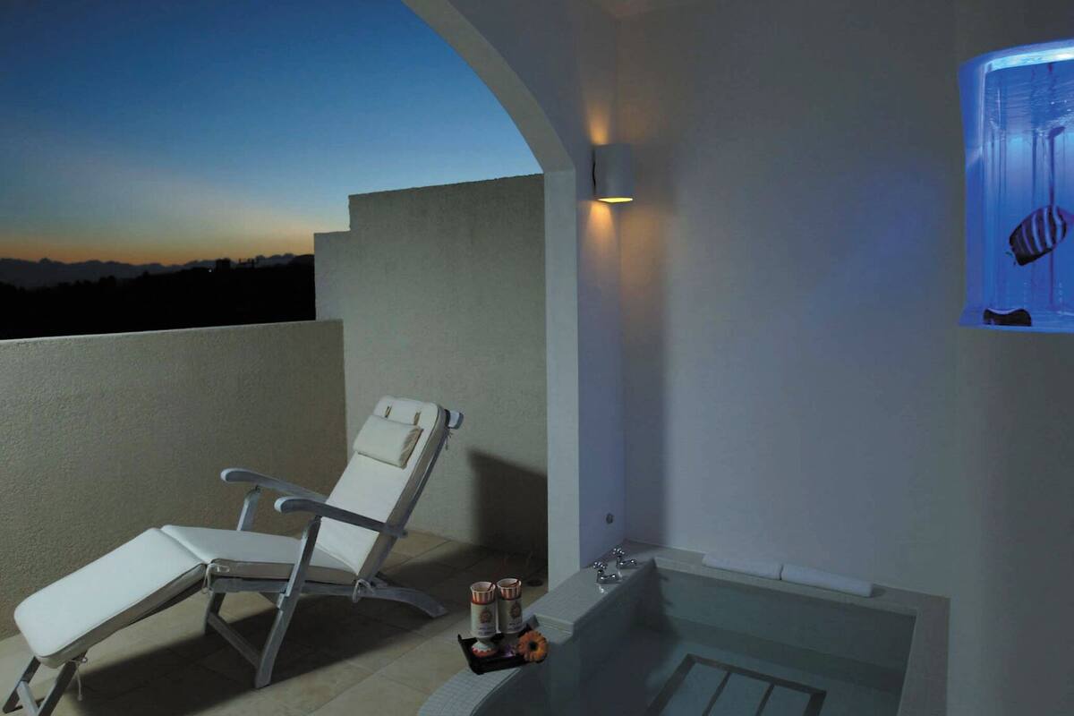 A balcony with a white lounge chair, a small table with two cups and a donut, and a wall-mounted aquarium.