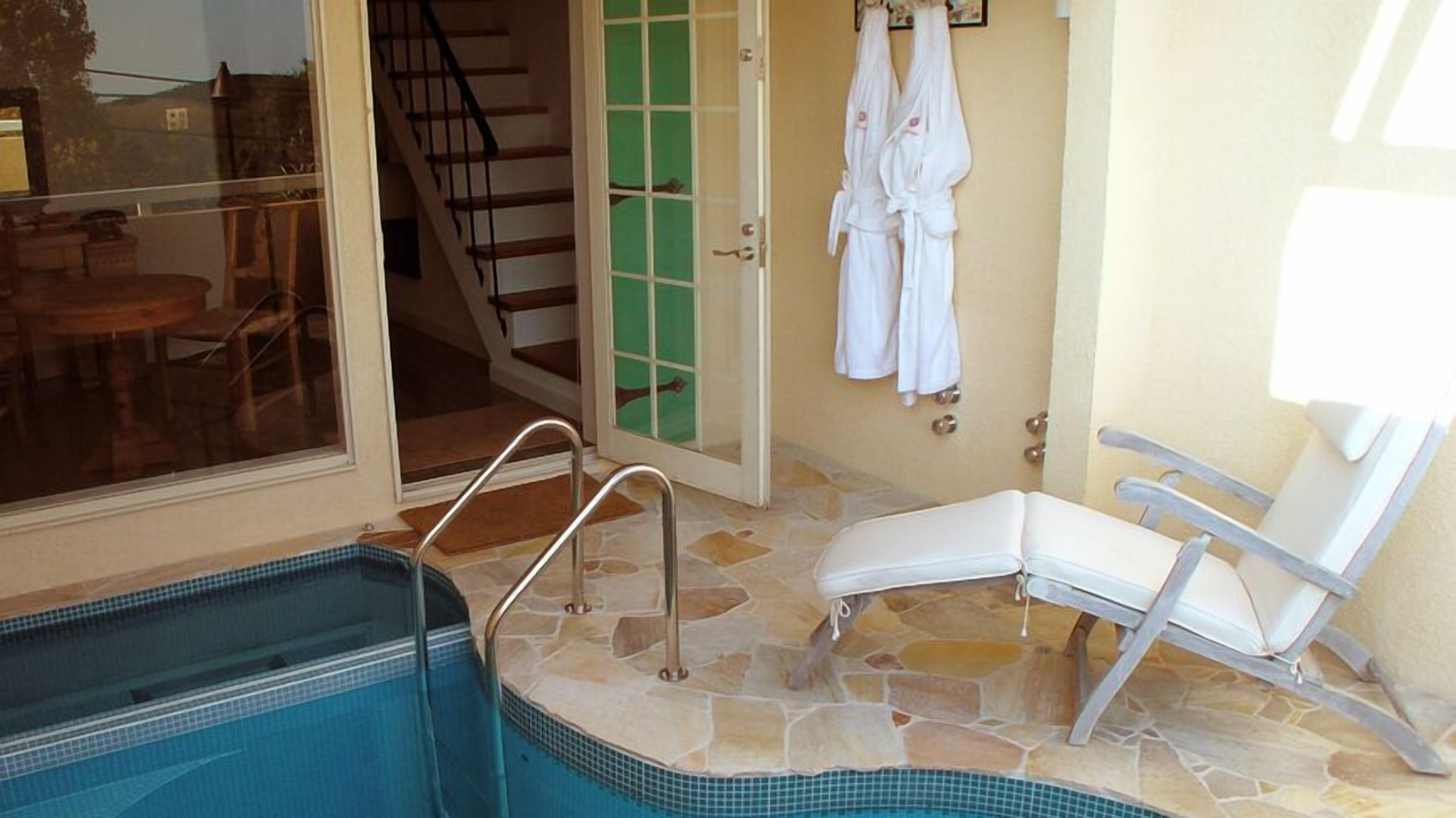 A pool area with a lounge chair, a glass door leading to a staircase, and a pair of white robes hanging on the wall.