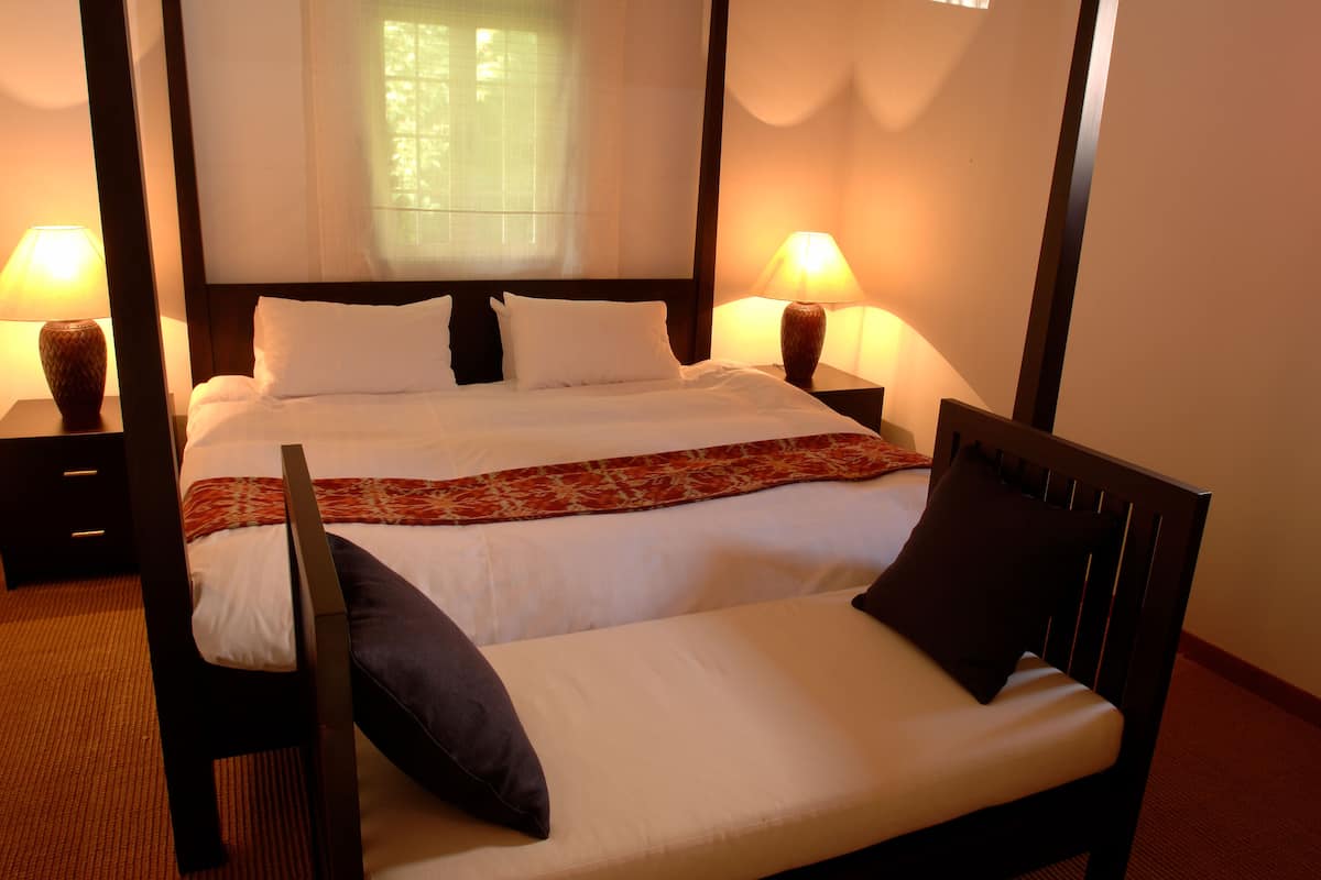 A four-poster bed with white linens, two bedside tables with lamps, and a view of greenery through the window.