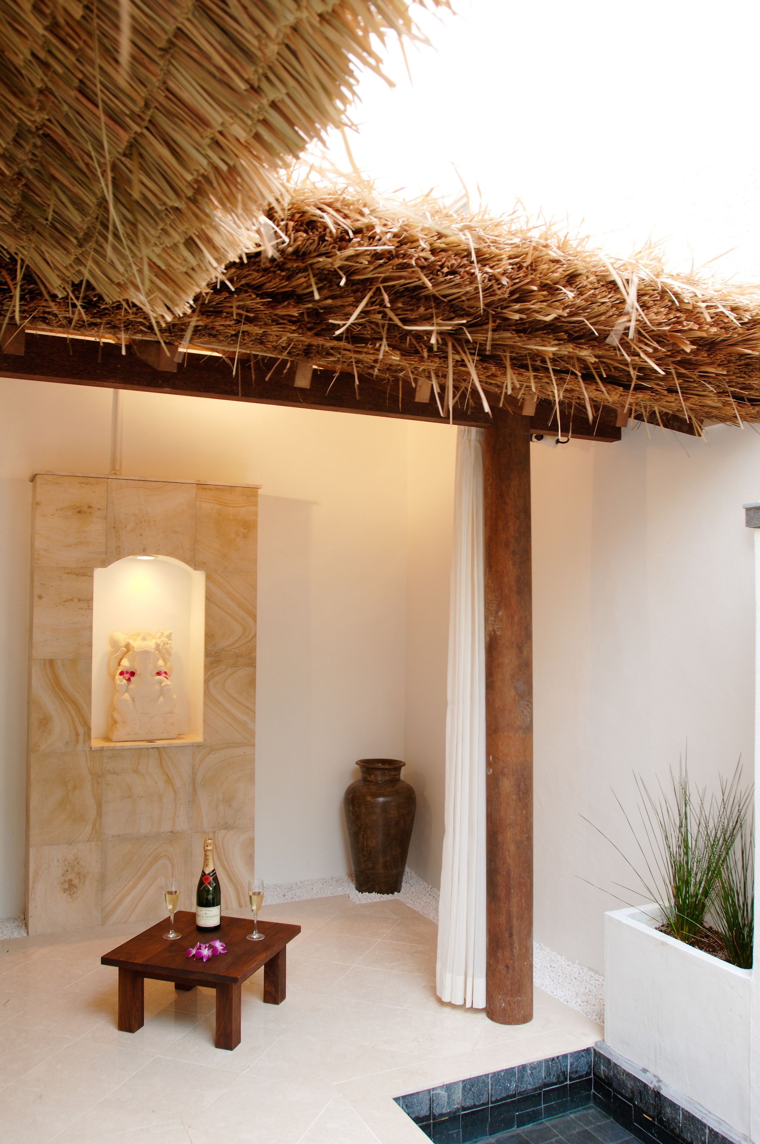 A small wooden table with a bottle and two glasses, a large vase, and a potted plant in a spa-like setting.
